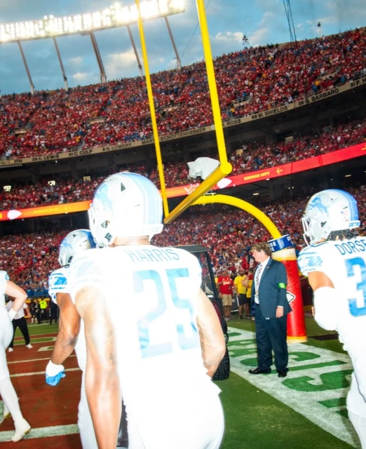 American football players in white and blue uniforms on a field with a large crowd in the background, football goalposts are visible.