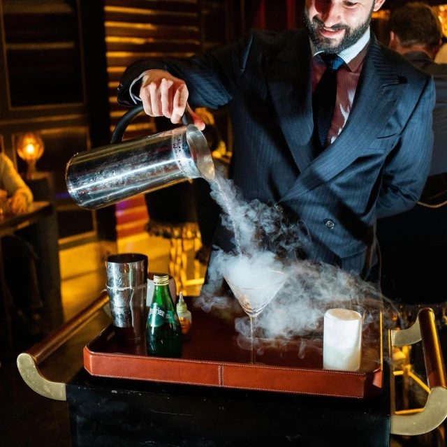 Bartender in a suit pours a smoking cocktail into a martini glass on a serving tray in a bar with moody lighting and glassware.