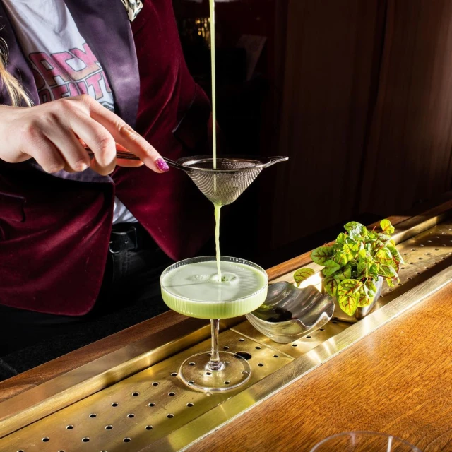 Professional bartender strains a bright green cocktail into a stemmed glass at a wood bar with decorative metal accents, indoors.