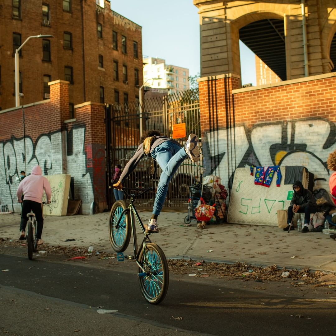 Adult riding bicycle on one wheel on paved street, with another cyclist, graffiti, and building in background during daytime.