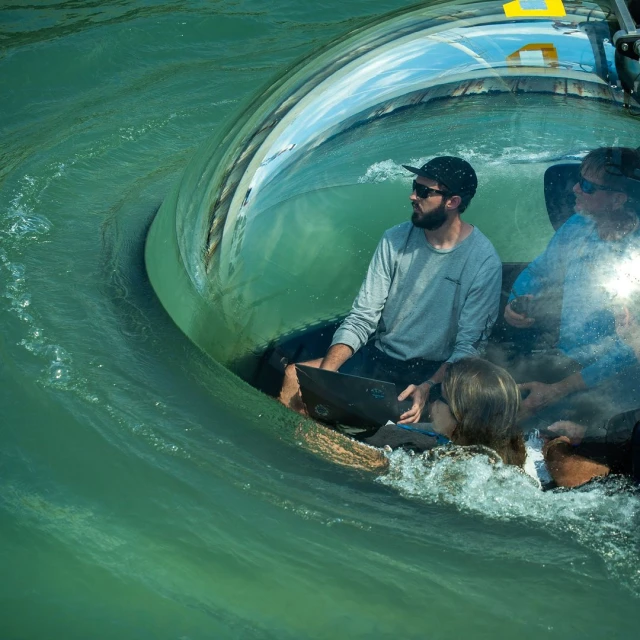 Three people inside a transparent submarine-like vessel in wavy green water, one using a laptop. An eye-level shot of the interior.