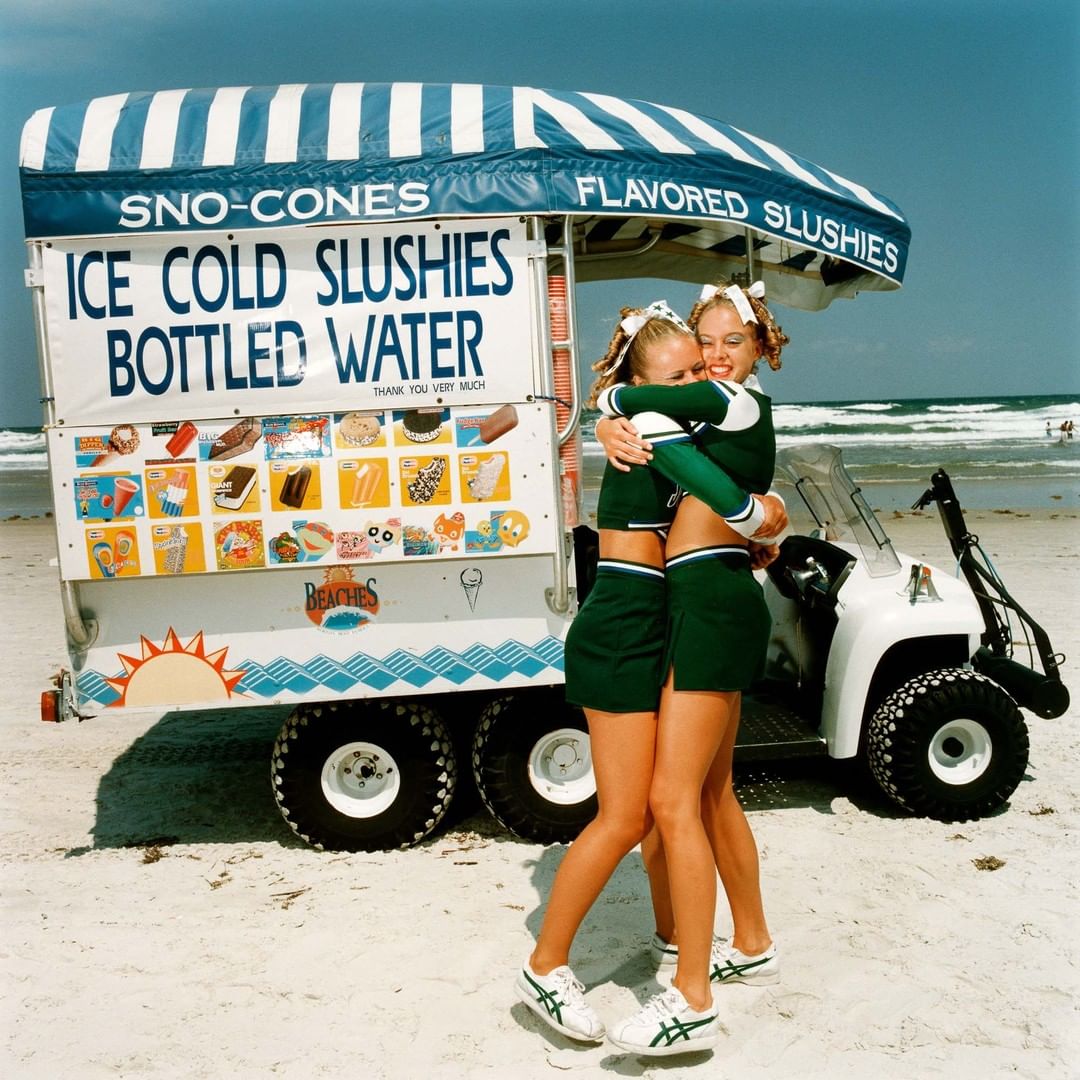 Two cheerleaders embrace near an ice cream cart on a sunny beach with ocean waves in the background, creating a playful summertime scene.