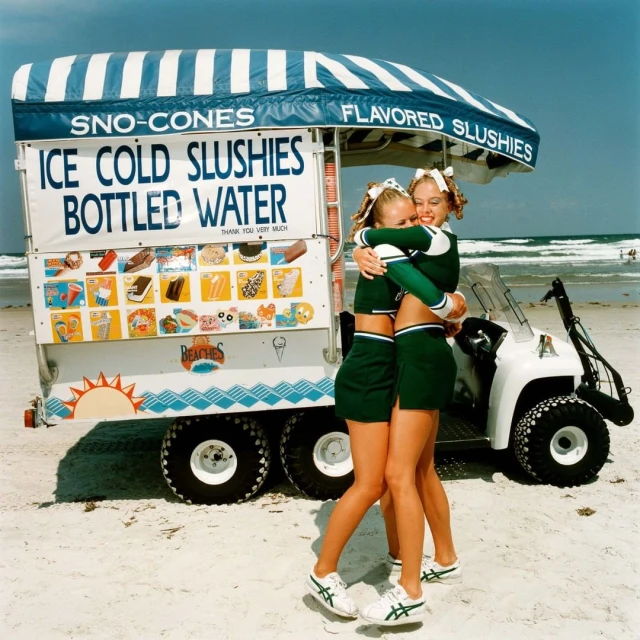 Two cheerleaders embrace near an ice cream cart on a sunny beach with ocean waves in the background, creating a playful summertime scene.