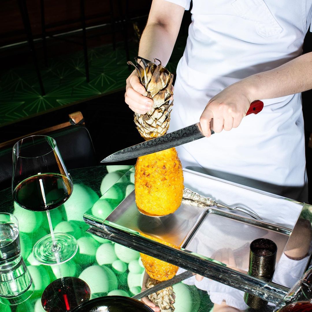 Chef cuts pineapple on a tray with a knife; a wine glass sits on a table with decorative green orbs in an indoor environment.