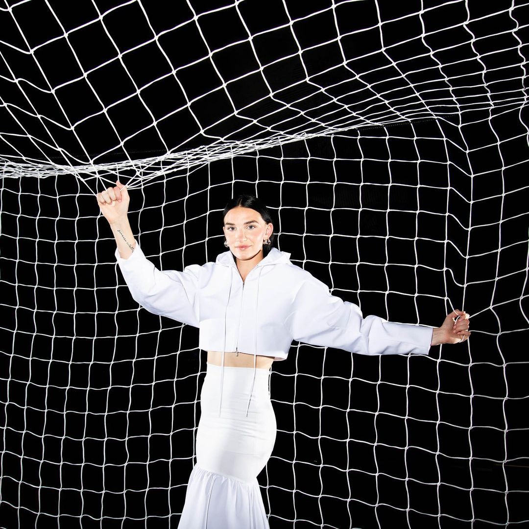 Woman in white outfit poses in front of a soccer net in a studio with black background, showcasing fashion and athletic elements.