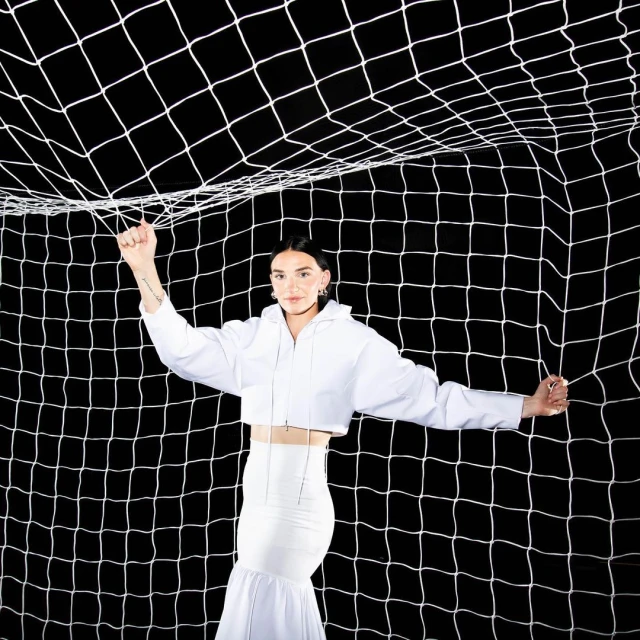 Woman in white outfit poses in front of a soccer net in a studio with black background, showcasing fashion and athletic elements.