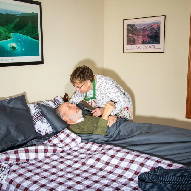 Elderly woman standing next to an elderly man in bed with a red-and-white checkered blanket. Pictures are on the wall.