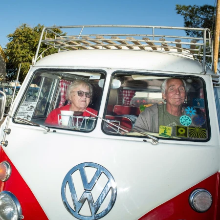 Elderly couple in a red and white Volkswagen van on a sunny day. The vintage van is parked outdoors, with trees visible in the background.