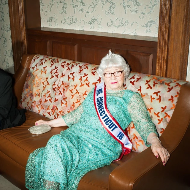 Elderly woman in a turquoise dress and tiara sits on an orange couch, wearing a 'Ms. Connecticut' sash, holding a clutch, indoor setting.