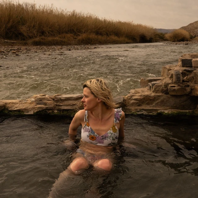 Woman in floral swimsuit sits in natural hot springs near river with reeds, enjoying the scenic view on an overcast day.