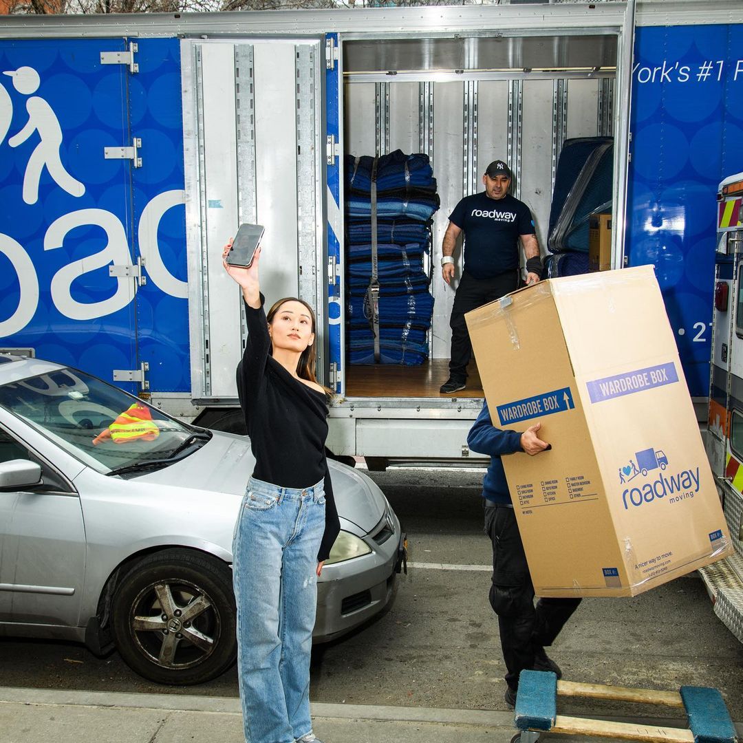 Woman takes a photo near a moving truck as movers handle a wardrobe box. A car is parked beside the truck on an urban street.