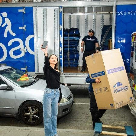 Woman takes a photo near a moving truck as movers handle a wardrobe box. A car is parked beside the truck on an urban street.