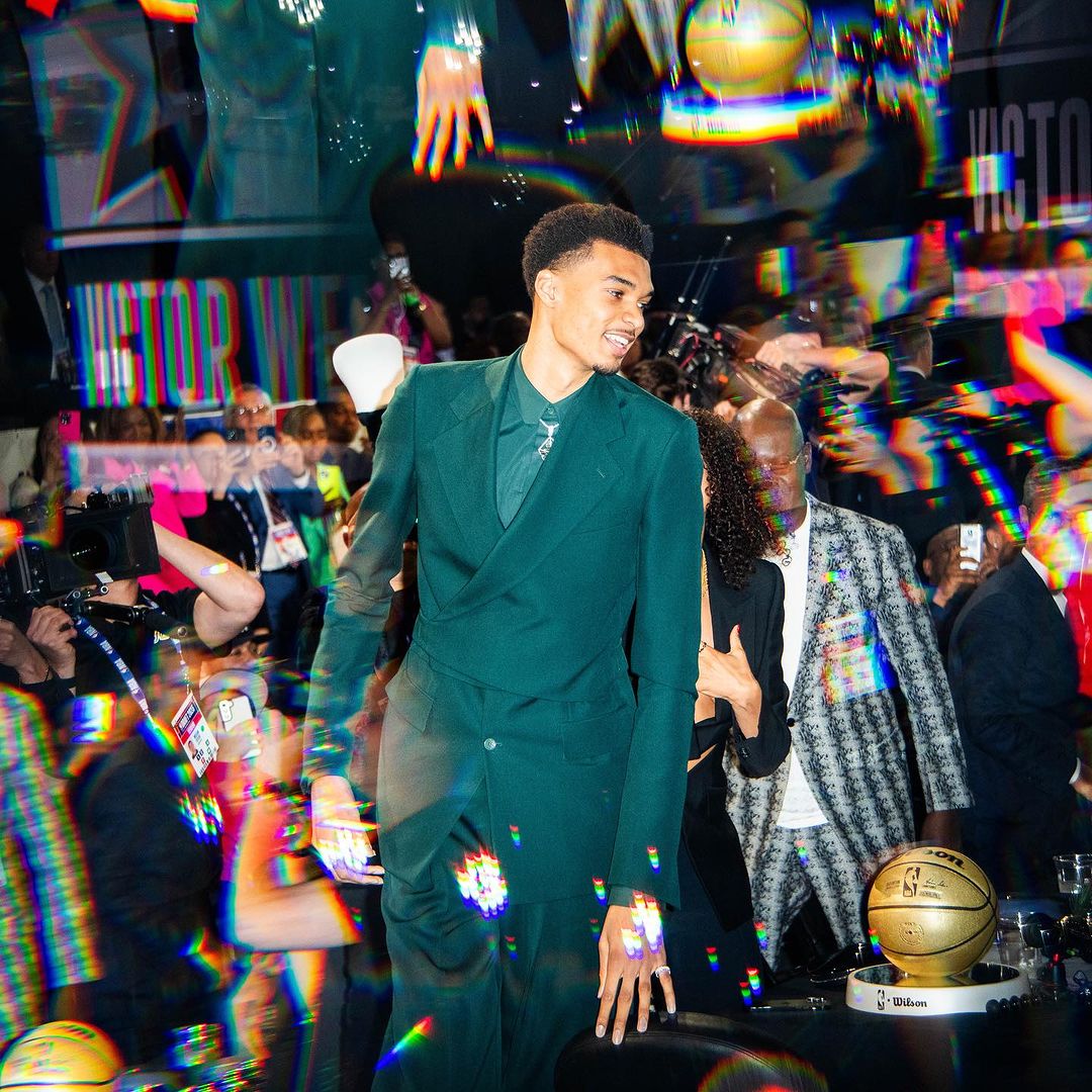 NBA prospect Victor Wembanyama poses in a green suit with a basketball trophy among a crowd and camera flashes.
