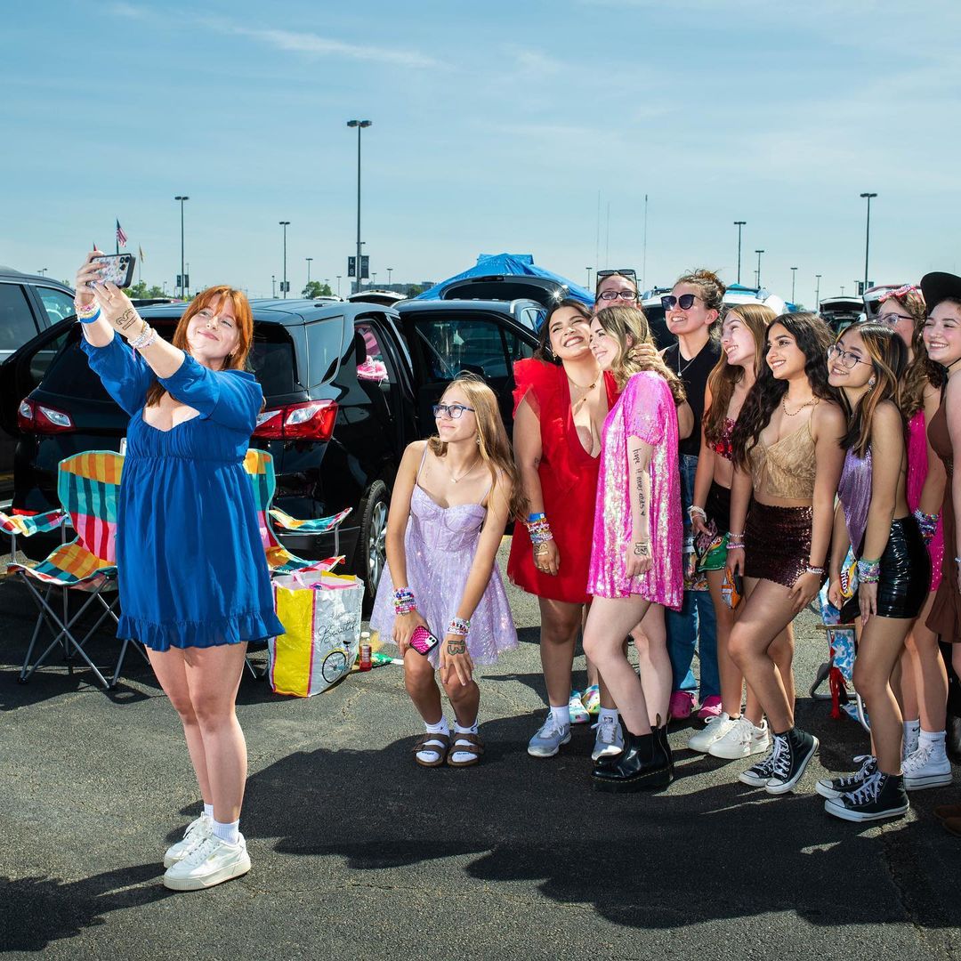 A group of young women are posing for a picture together in a sunny outdoor setting, dressed in colourful outfits with cars in the background.