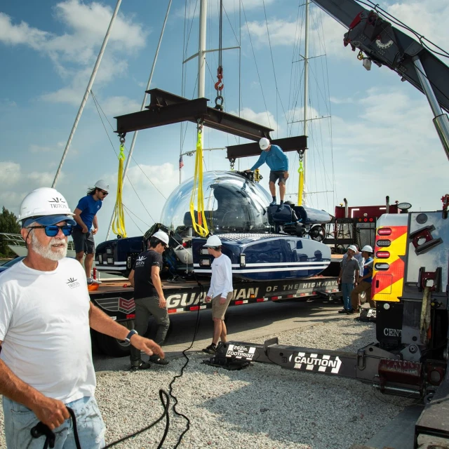 Submersible is lifted by crane with construction workers in hard hats on sunny day, blue sky with clouds, outdoor scene.