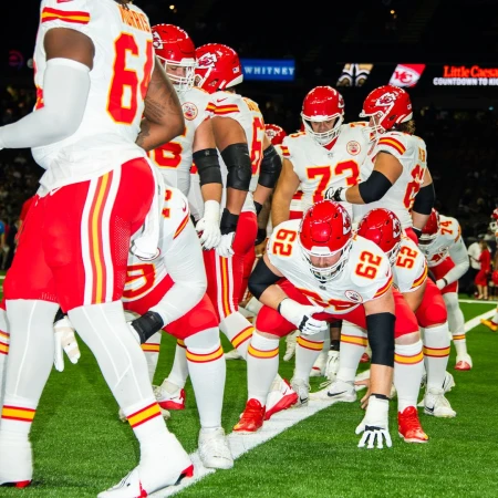 American football players in white and red uniforms line up on a green field in an indoor stadium, preparing for a play.
