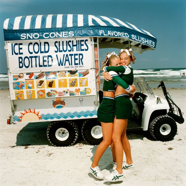 Two cheerleaders hugging in front of a snow cone and slushie stand on a sunny beach with the ocean in the background.
