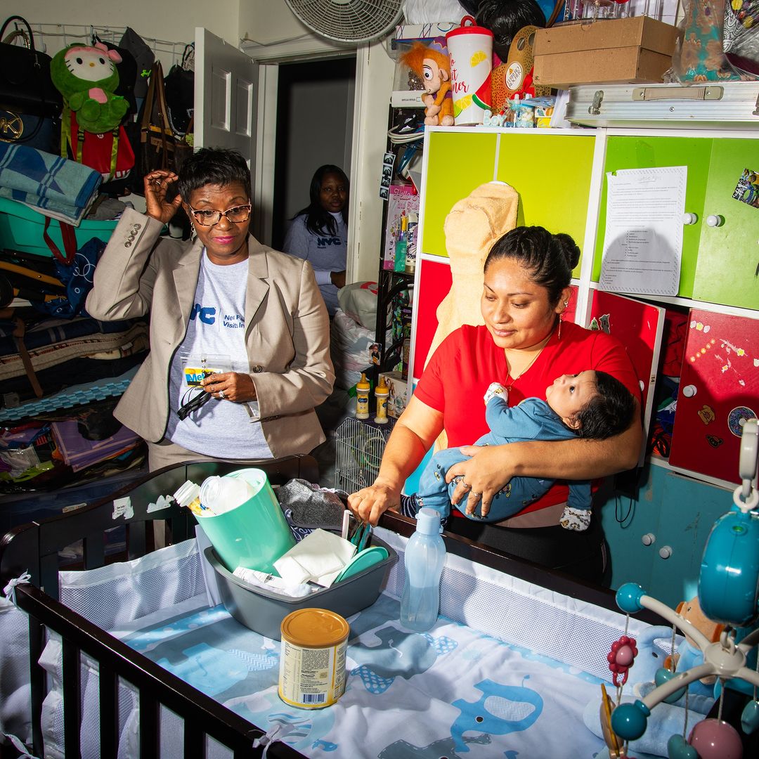 A woman holds a baby near a crib as a social worker stands nearby in a cluttered home, offering support and assistance.