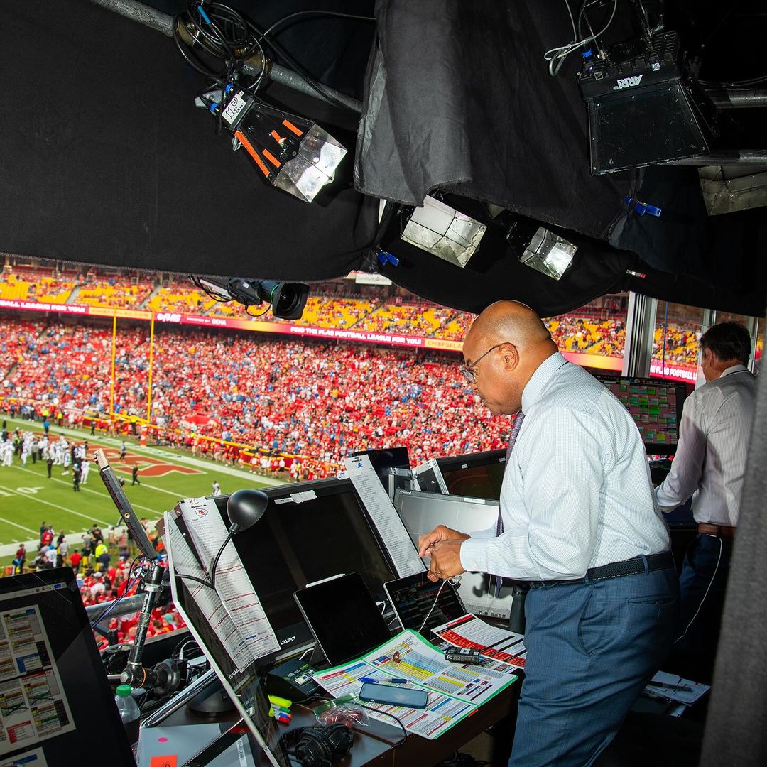 View from a press box overlooking an American football game. A man in a white shirt works behind a console with a stadium visible beyond.