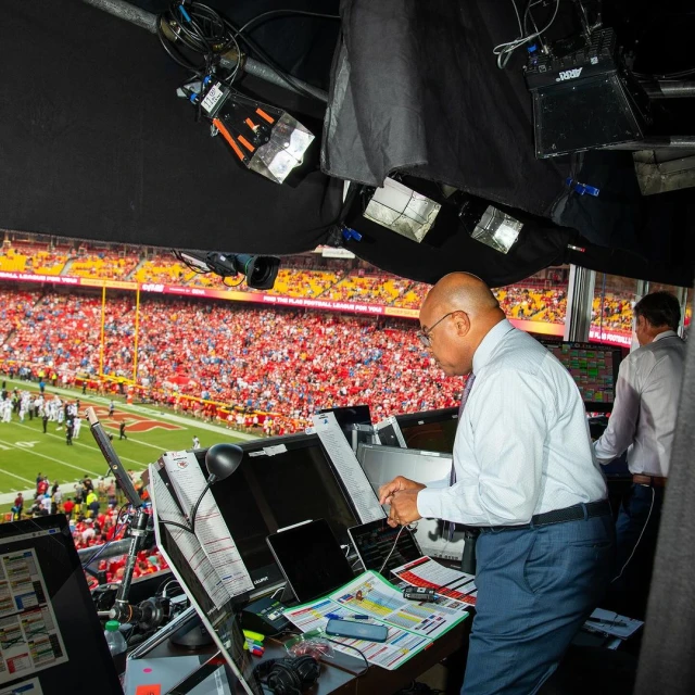 View from a press box overlooking an American football game. A man in a white shirt works behind a console with a stadium visible beyond.