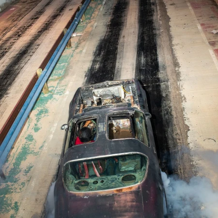 Overhead shot of a damaged black car with a driver on a racetrack with tire tracks, smoke billowing from the car, and blue rails on the sides.