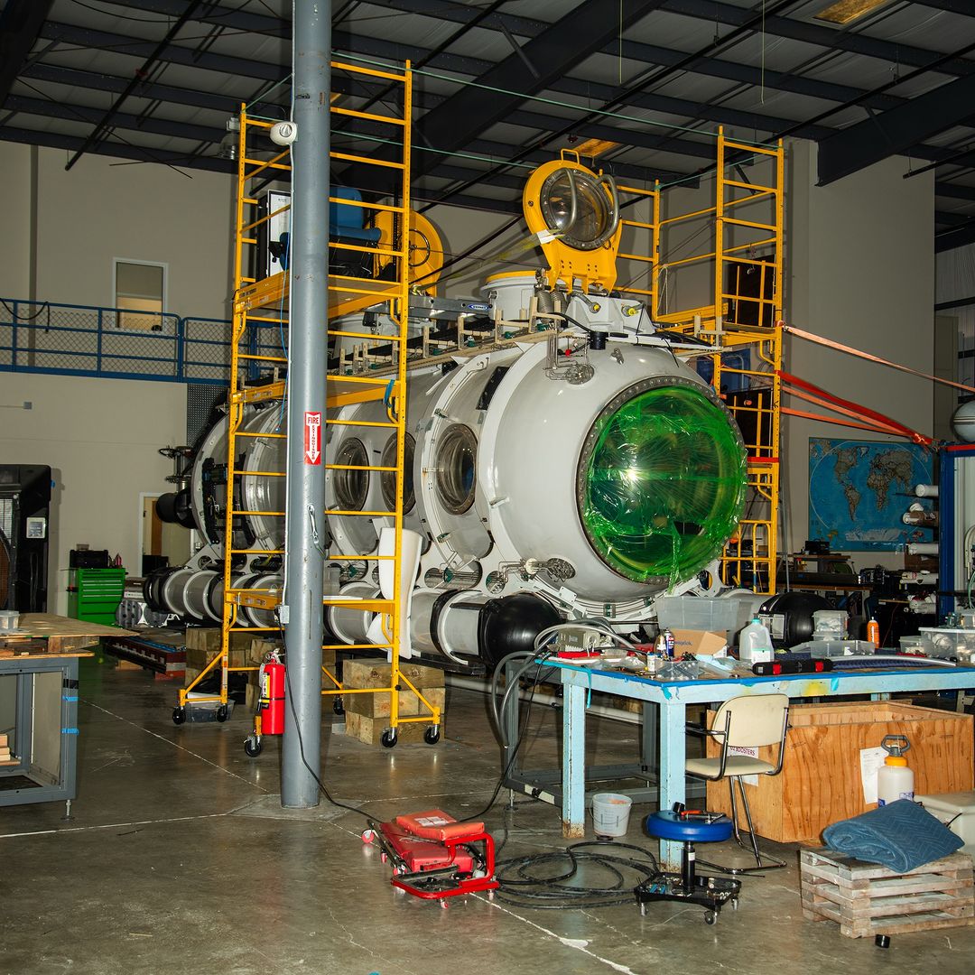 Large white submarine is surrounded by yellow scaffolding in a warehouse setting, with tools and equipment nearby, and a map on the wall.
