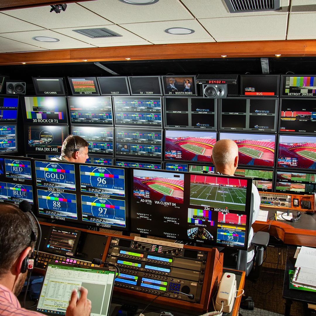 Inside a television control room, three people monitor numerous screens displaying live sports and other content, adjusting audio and video.