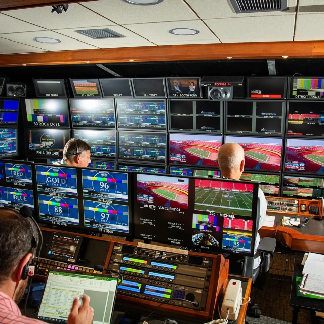 Inside a television control room, three people monitor numerous screens displaying live sports and other content, adjusting audio and video.