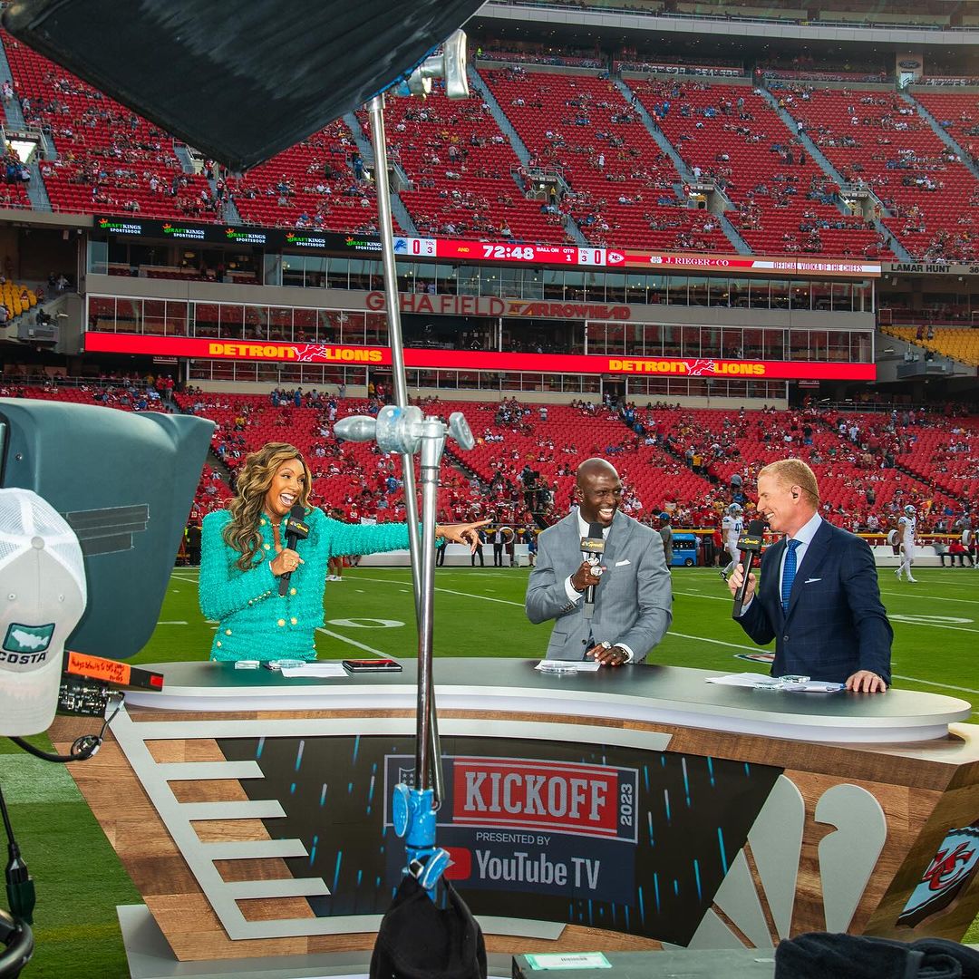 Broadcast of football kickoff show in stadium with three commentators at desk, green field, audience, and broadcast lighting present.