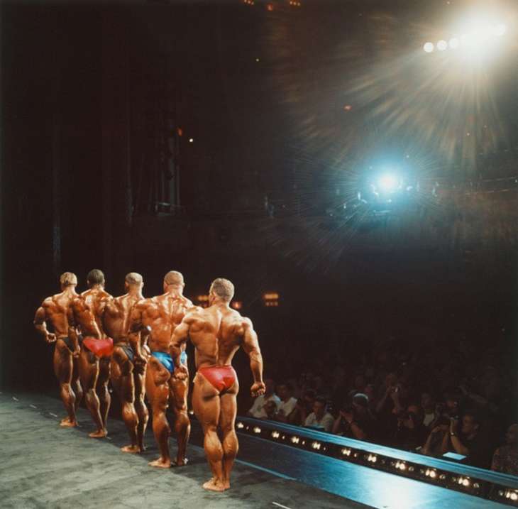A line of muscular bodybuilders in posing trunks are displayed from the back on a stage, facing a dark crowd and bright stage lights.