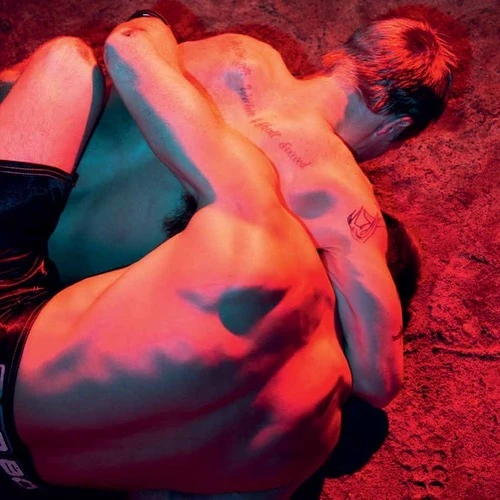 Two shirtless adult male wrestlers grappling on a textured surface, lit with red and blue gels, showcasing physical exertion and body art.