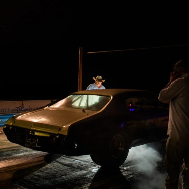 Vintage muscle car burnout at night with crowd watching. Rear view of classic black automobile smoking tires at drag strip.