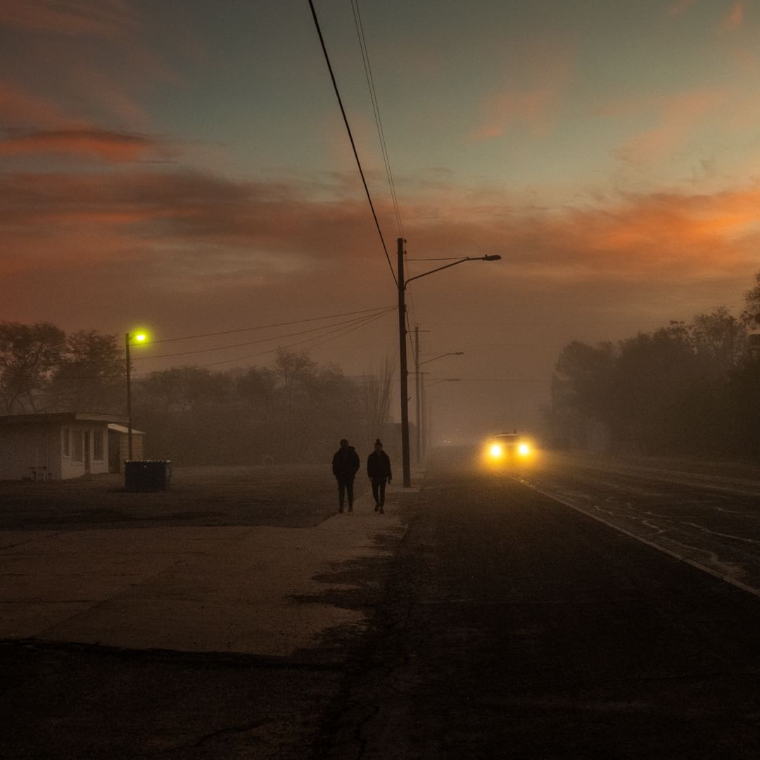 Two silhouetted figures walk along a foggy road beside railroad tracks as a car's headlights approach in the distance at dusk.