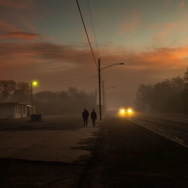 Two silhouetted figures walk along a foggy road beside railroad tracks as a car's headlights approach in the distance at dusk.