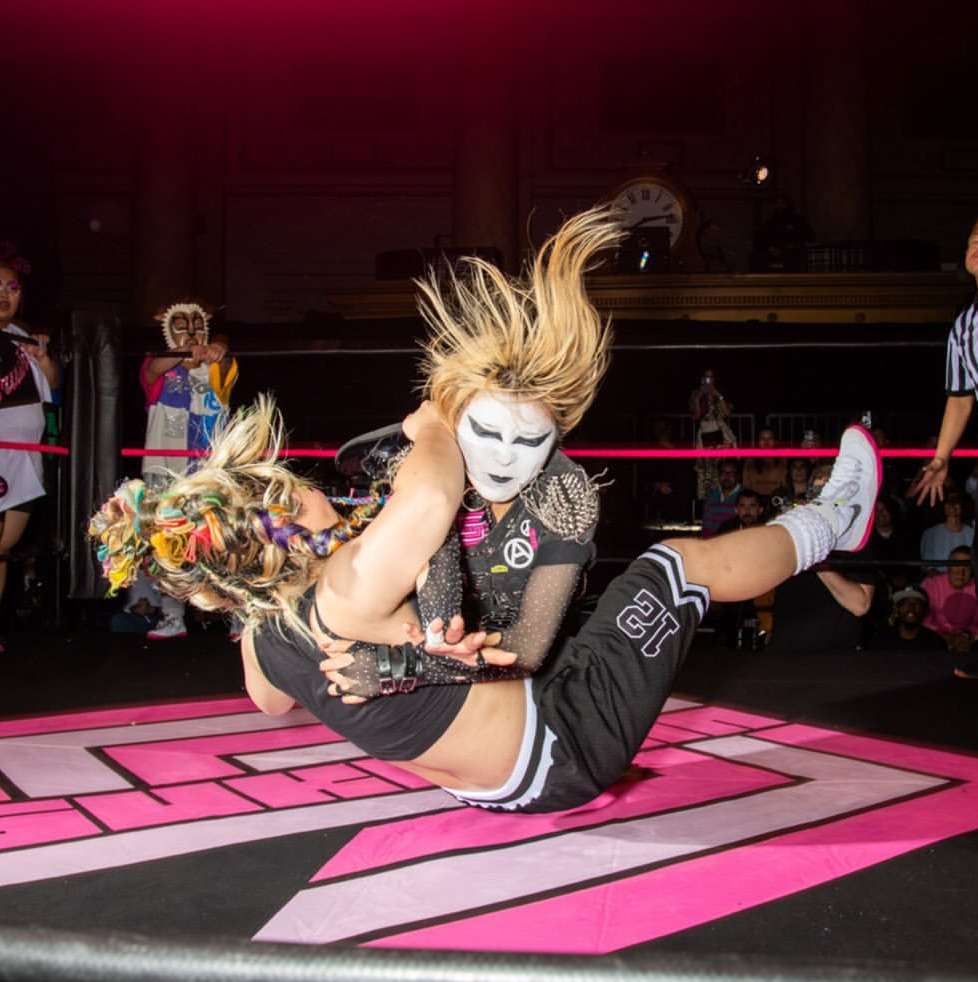 Two female wrestlers in a wrestling ring during a match. One wrestler in a headlock with blonde hair, face paint, and athletic attire.