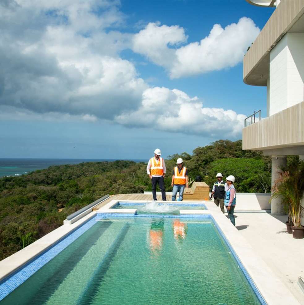 Exterior view of a modern home with a blue-tiled swimming pool and four construction workers wearing vests and hard hats.