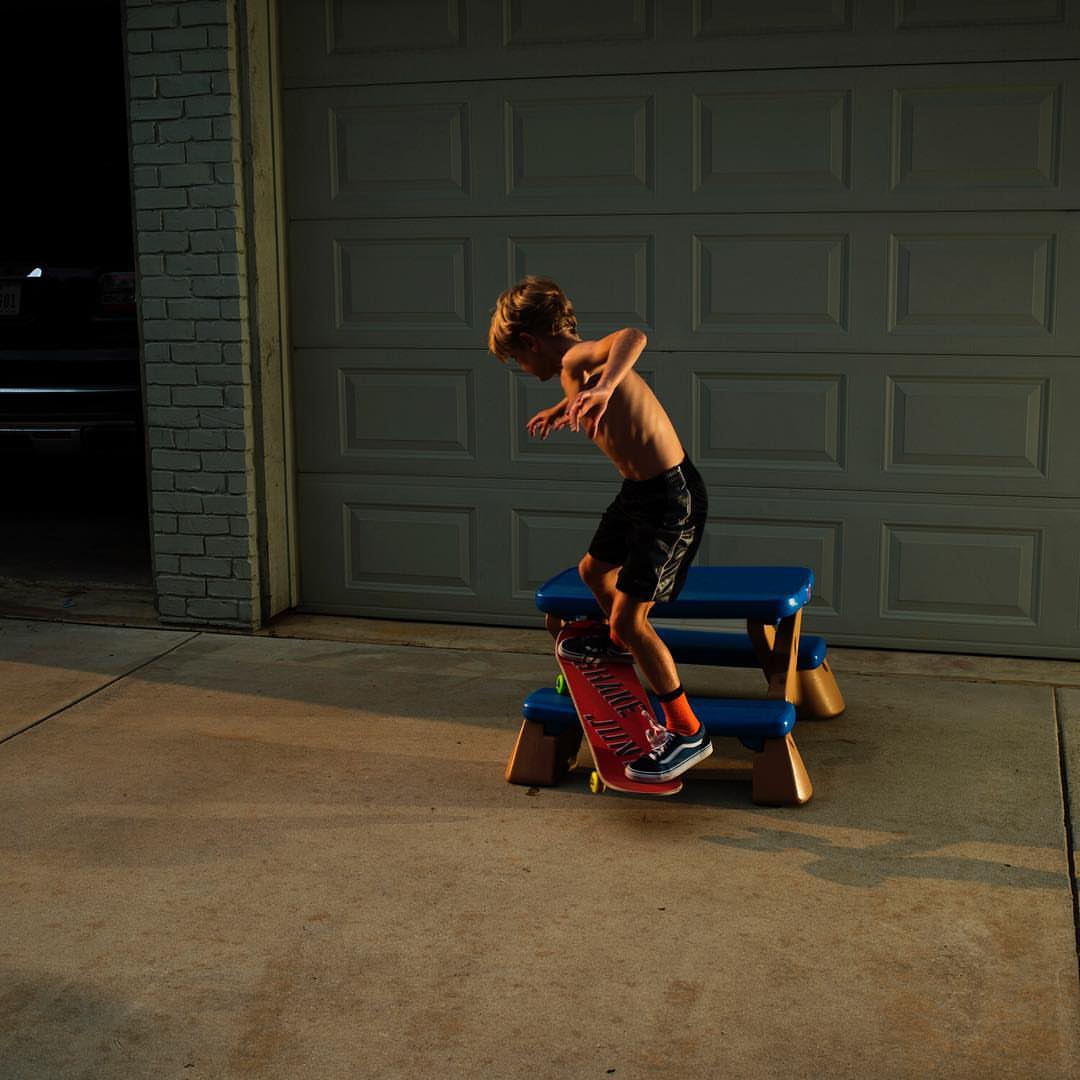 Boy on skateboard stands on a blue step stool in a driveway in front of a garage door, lit by overhead light.