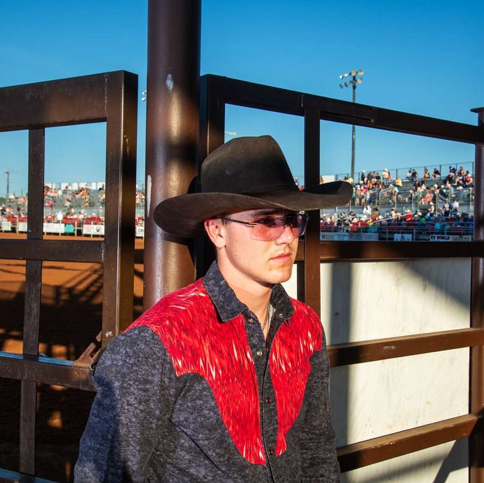 Young man in cowboy hat and red-detailed shirt at rodeo arena. Bright sunlight, daytime shot of a person standing outdoors at an event.