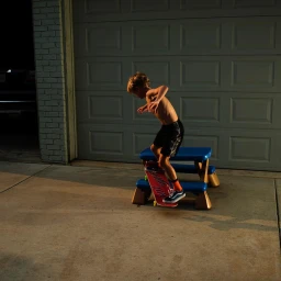Young boy skateboarding on a small blue picnic bench outside a closed garage door, captured by Brian Finke