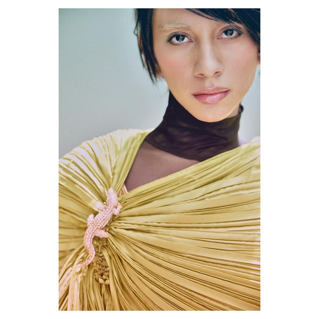 Portrait of a person with light hair and a yellow pleated dress, featuring a pink lizard brooch, against a soft blue background.