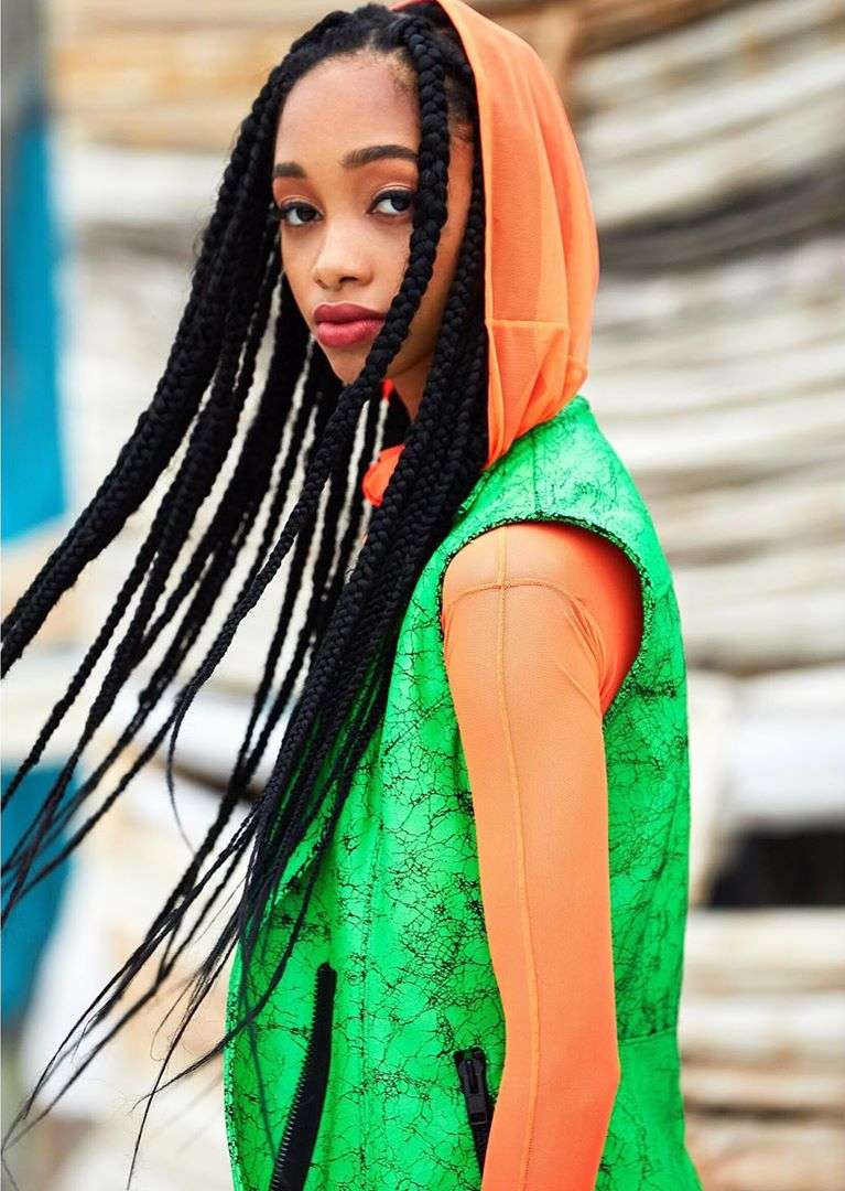 Young woman in a green vest and orange hooded top, looking at the camera with braids and red lipstick, outdoors, fashion-forward and stylish.
