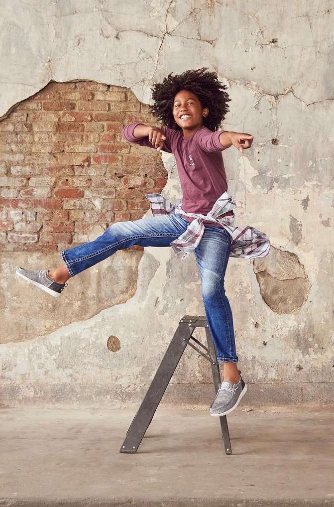 Energetic young boy strikes dynamic pose on ladder against weathered wall, wearing jeans, tee, and shoes, radiating joy and movement.