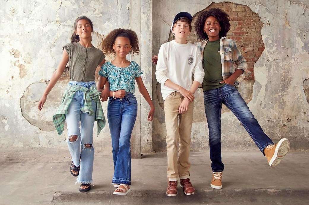 Four young teens pose in fashionable outfits against a distressed wall. Light blue, blue and green colors are dominant.