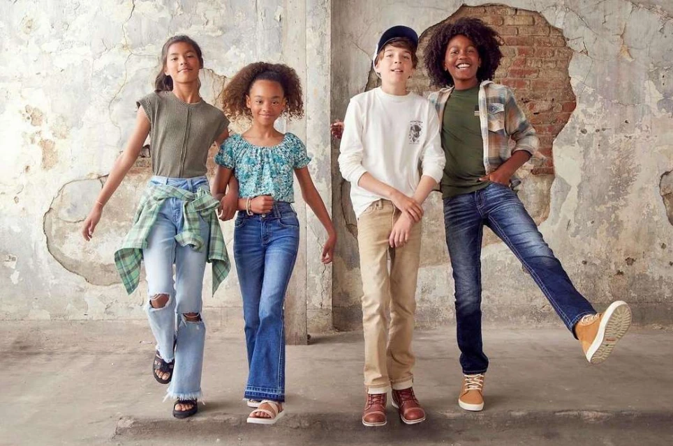 Four young teens pose in fashionable outfits against a distressed wall. Light blue, blue and green colors are dominant.