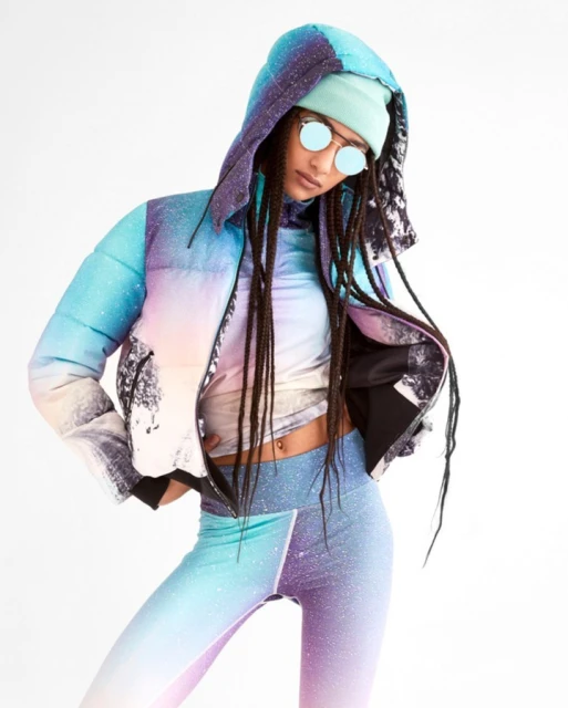 Woman in galaxy print puffer jacket, leggings, and beanie poses in a photo studio against a white backdrop wearing round sunglasses.