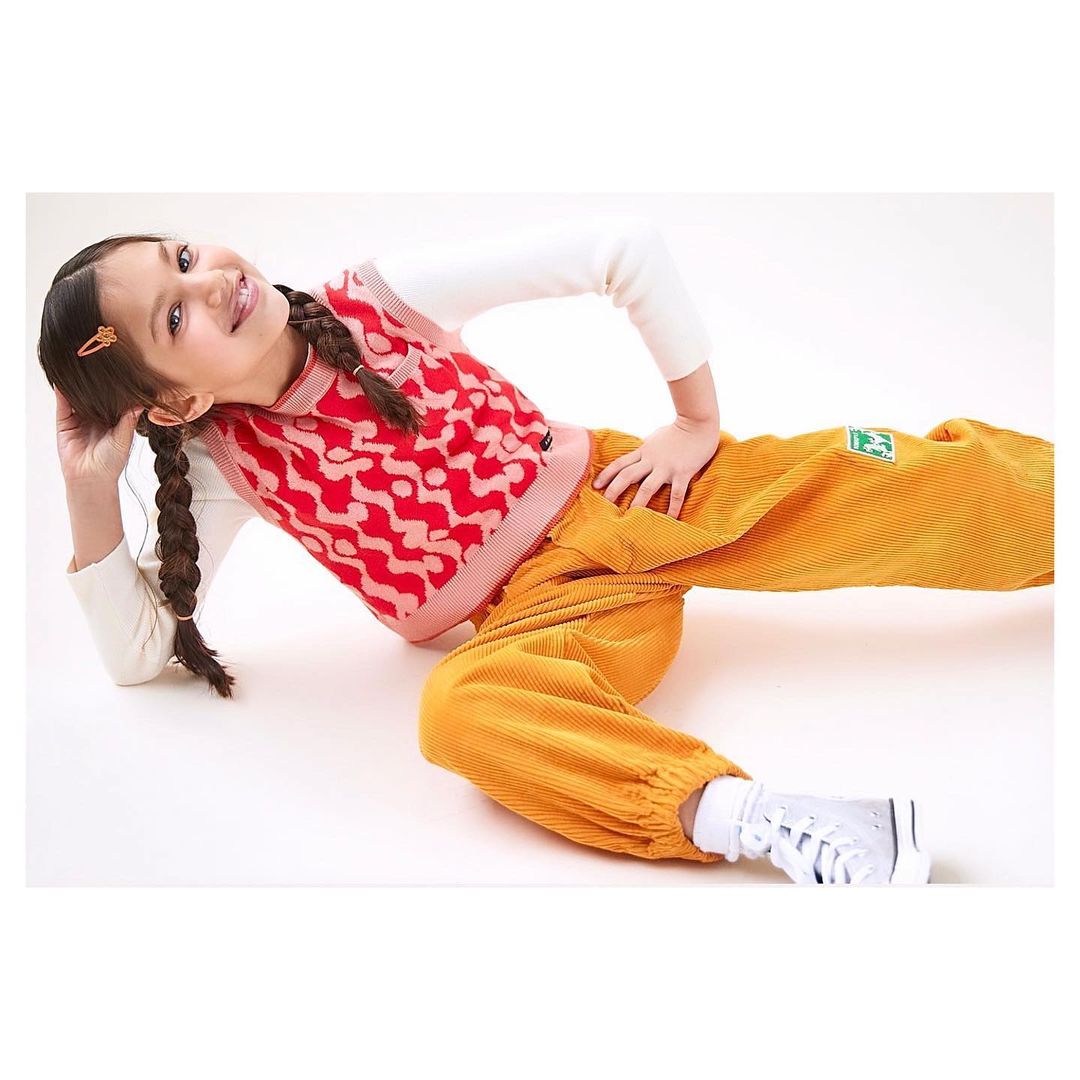 Smiling young girl in a patterned red vest, white long-sleeve shirt, and yellow corduroy pants, playfully posing on a white background.