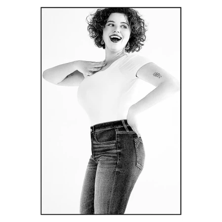 Black and white portrait of a woman in a white t-shirt and jeans, smiling with her hand on her chest, against a plain white background.
