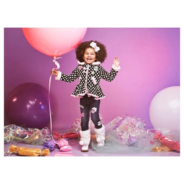 A cute child stands in front of a pink and purple background, holding a large pink balloon, wearing a polka-dotted coat and jeans.