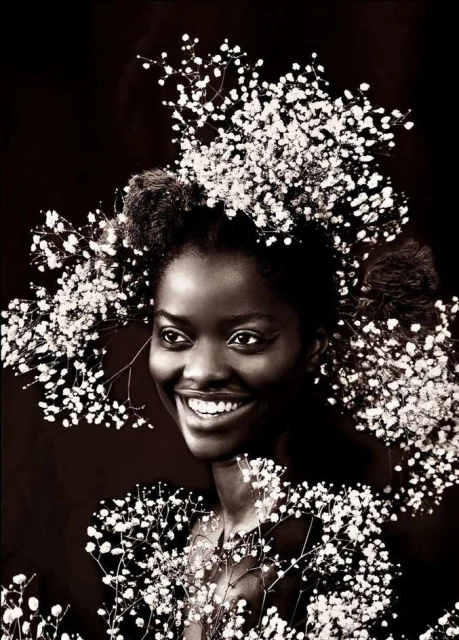 Close-up portrait of a smiling Black woman with an afro, adorned with baby's breath flowers around her head and shoulders against a dark background.