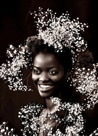 Close-up portrait of a smiling Black woman with an afro, adorned with baby's breath flowers around her head and shoulders against a dark background.